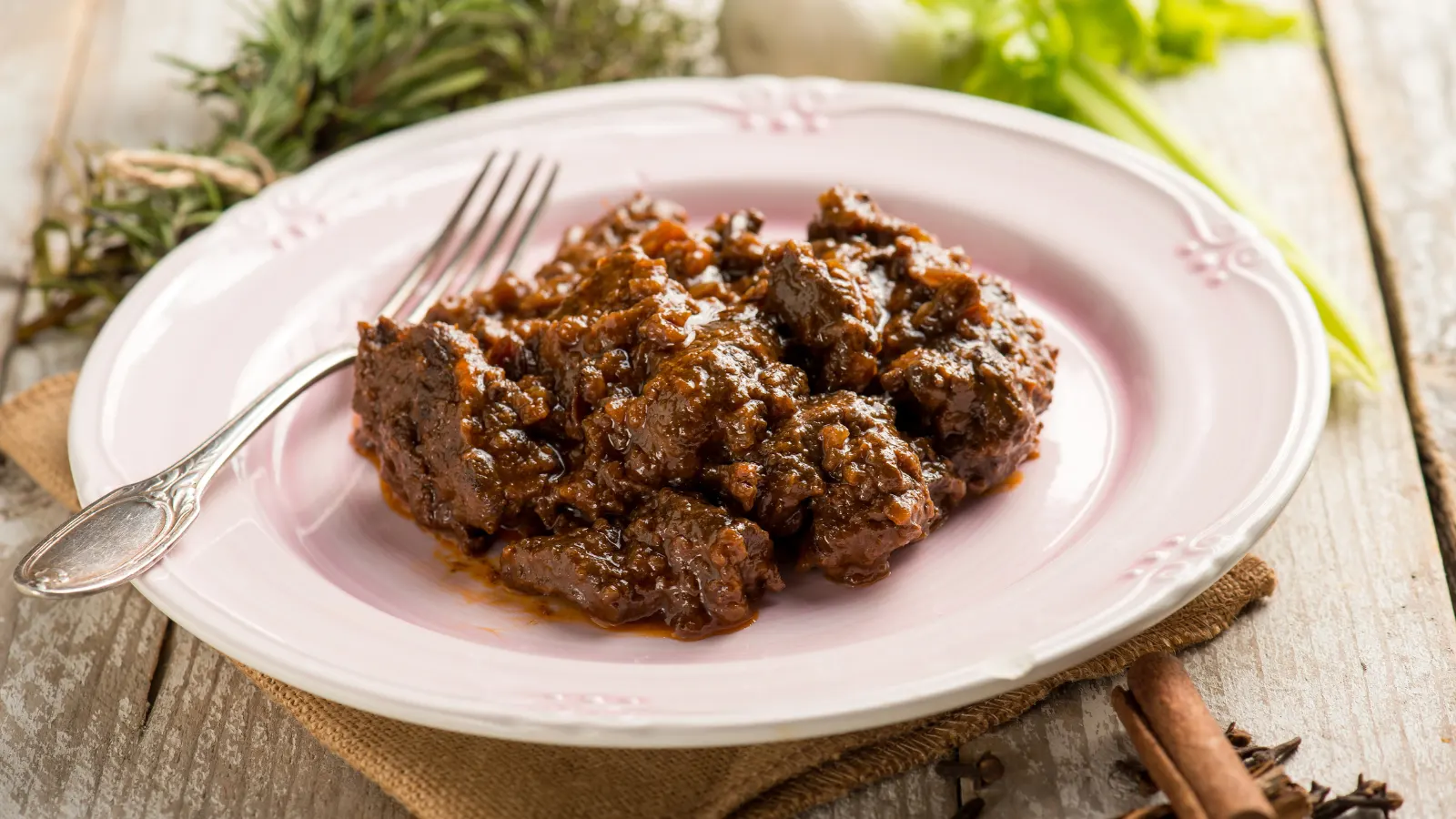Hirschgulasch: Rezept mit kräftiger Soße und Wacholder Hirschgulasch mit dunkler Soße auf einem Teller, daneben Kräuter und Zimt