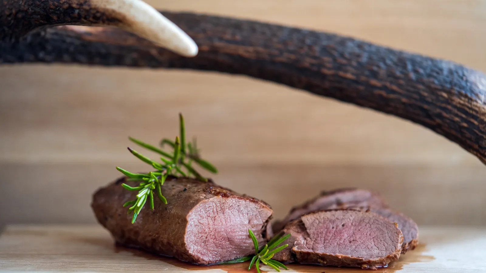 Rehrücken Rezept: rosa aus dem Ofen mit Wildsoße Rosa gegartes Wildfilet in Scheiben auf einem Holzbrett, im Hintergrund eine Geweihstange